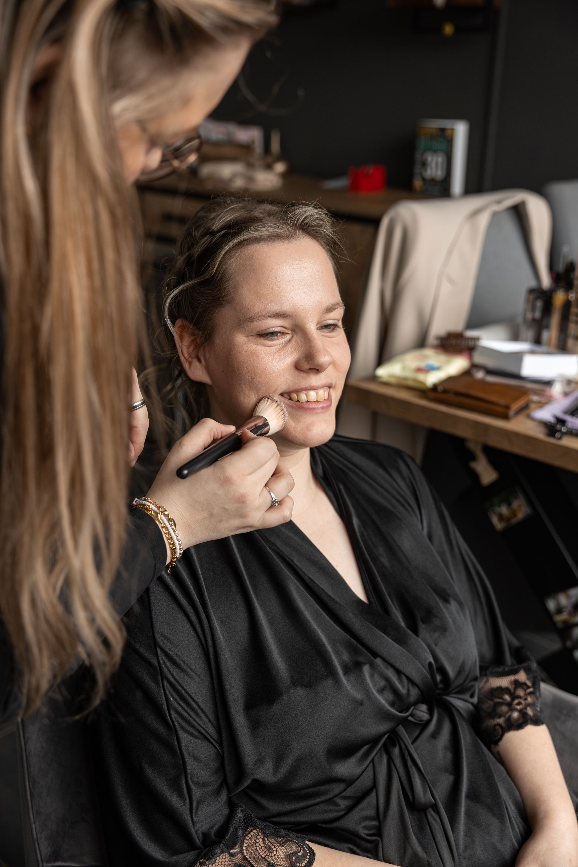 Vrouw die backstage wordt opgemaakt, lachend in een badjas. Goedkope bruidsfotografie. Bruiloft fotograaf Hellevoetsluis.