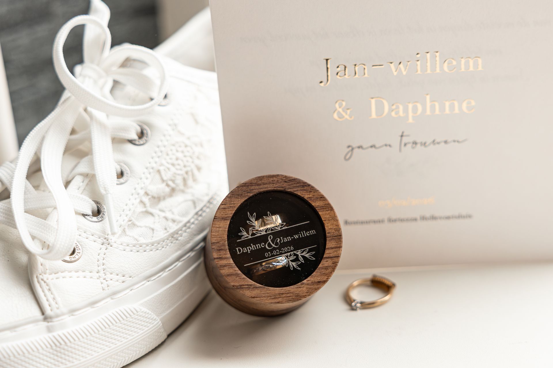 Witte sneakers naast een houten ringdoosje en een gouden ring, met uitnodiging voor bruiloft op de achtergrond.