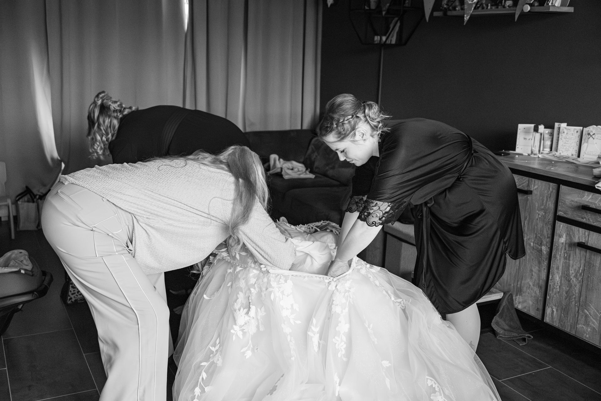 Twee mensen buigen zich over een jonge vrouw in een galajurk en trekken haar jurk recht in een kamer. Bruidsjurk fotografie
