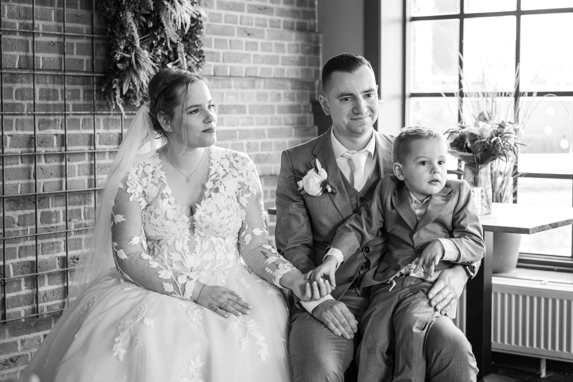 Zwart-wit trouwportret van een bruidsstel die bij een raam zitten, lachend en hand in hand. Bruidsfotograaf. Bruiloft foto's.