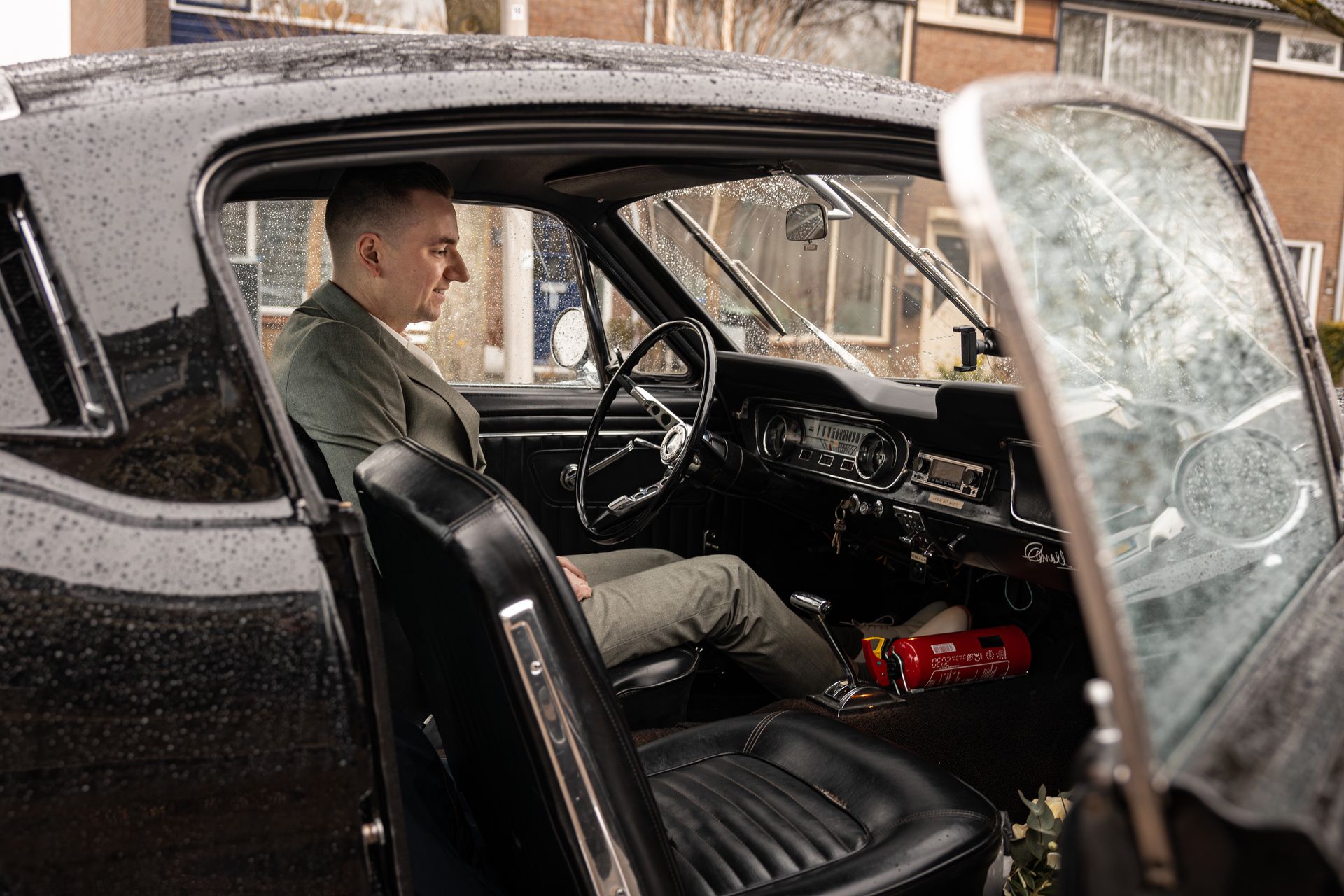 Een man zit achter het stuur van een zwarte oldtimer met de deur open. Bruidegom. Goedkope Bruidsfotografie Zuid Holland.