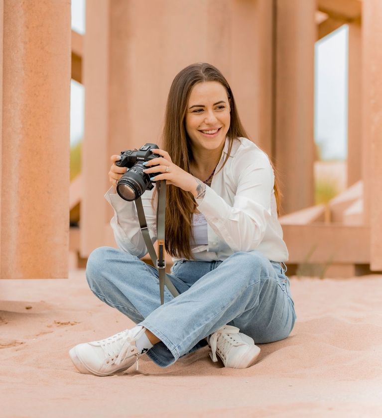 Vrouw zit op een muur, lachend terwijl ze een camera vasthoudt, met perzikkleurige pilaren. Fotograaf Zuidland.