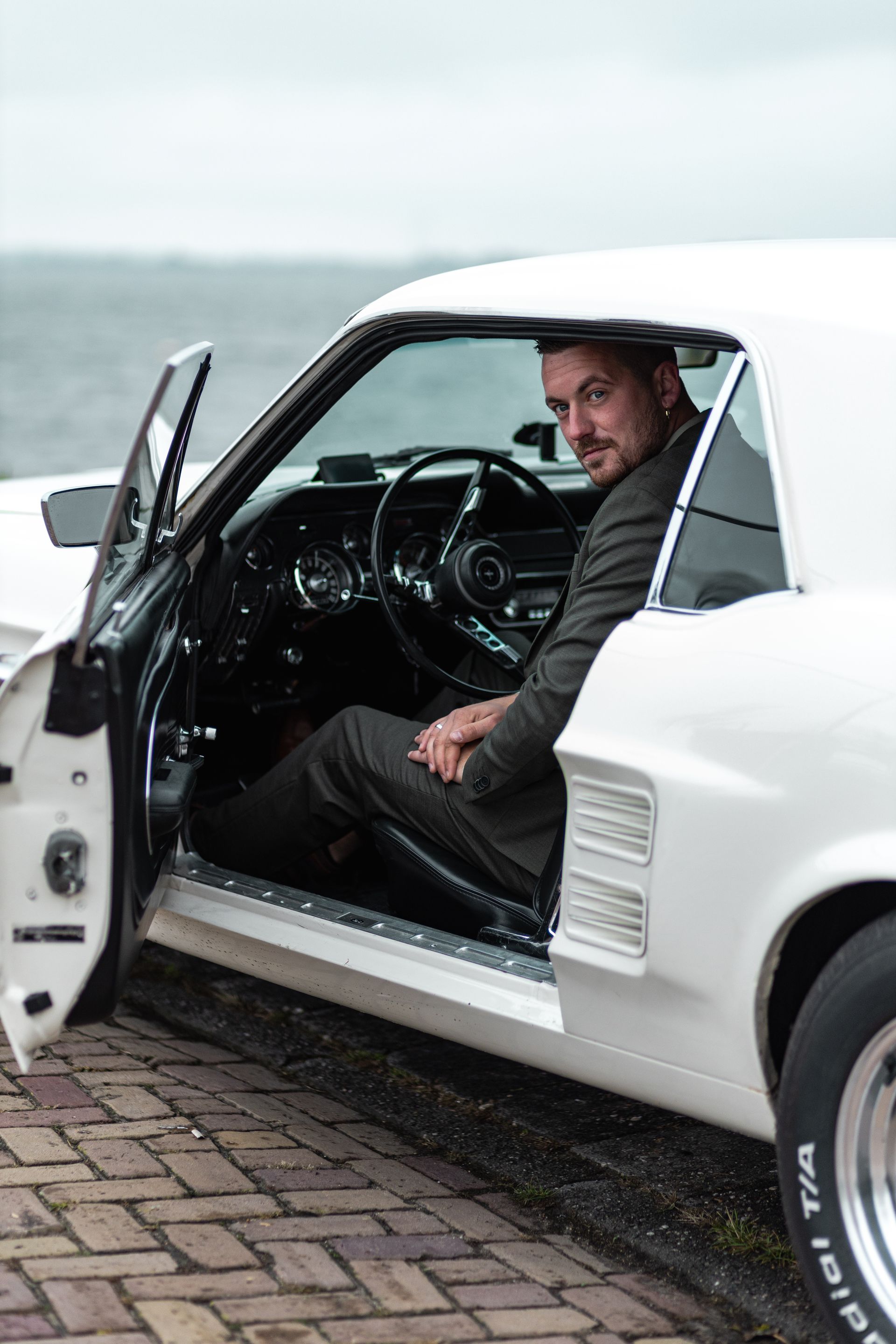 Een man zit in een witte klassieke auto met de deur open, geparkeerd aan de waterkant. Bruidsfotograaf Hellevoetsluis.