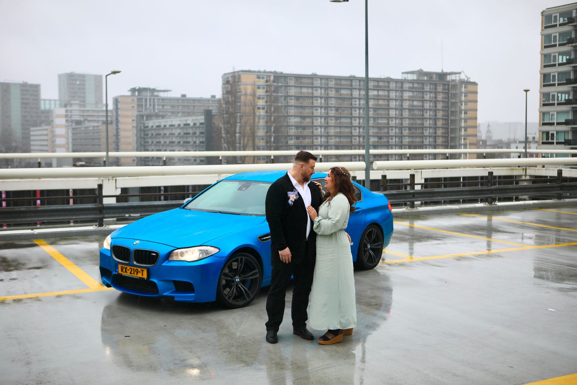 Een stel staat naast een felblauwe auto op een regenachtig dak met stadsgezichten op de achtergrond.