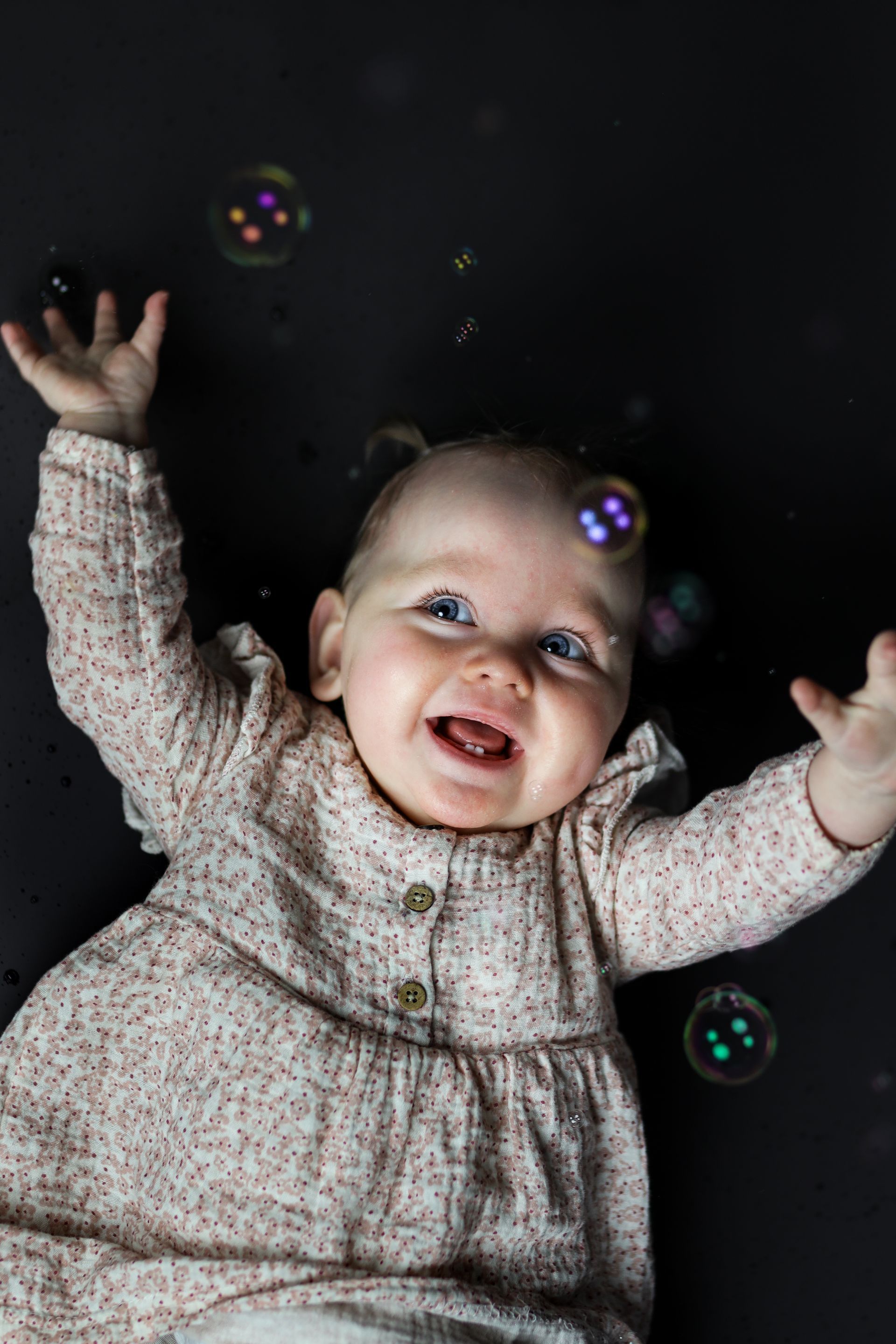 Een lachende baby in een grijze jurk met opgeheven armen reikt naar zwevende bubbels. Kinderfotografie in de studio.