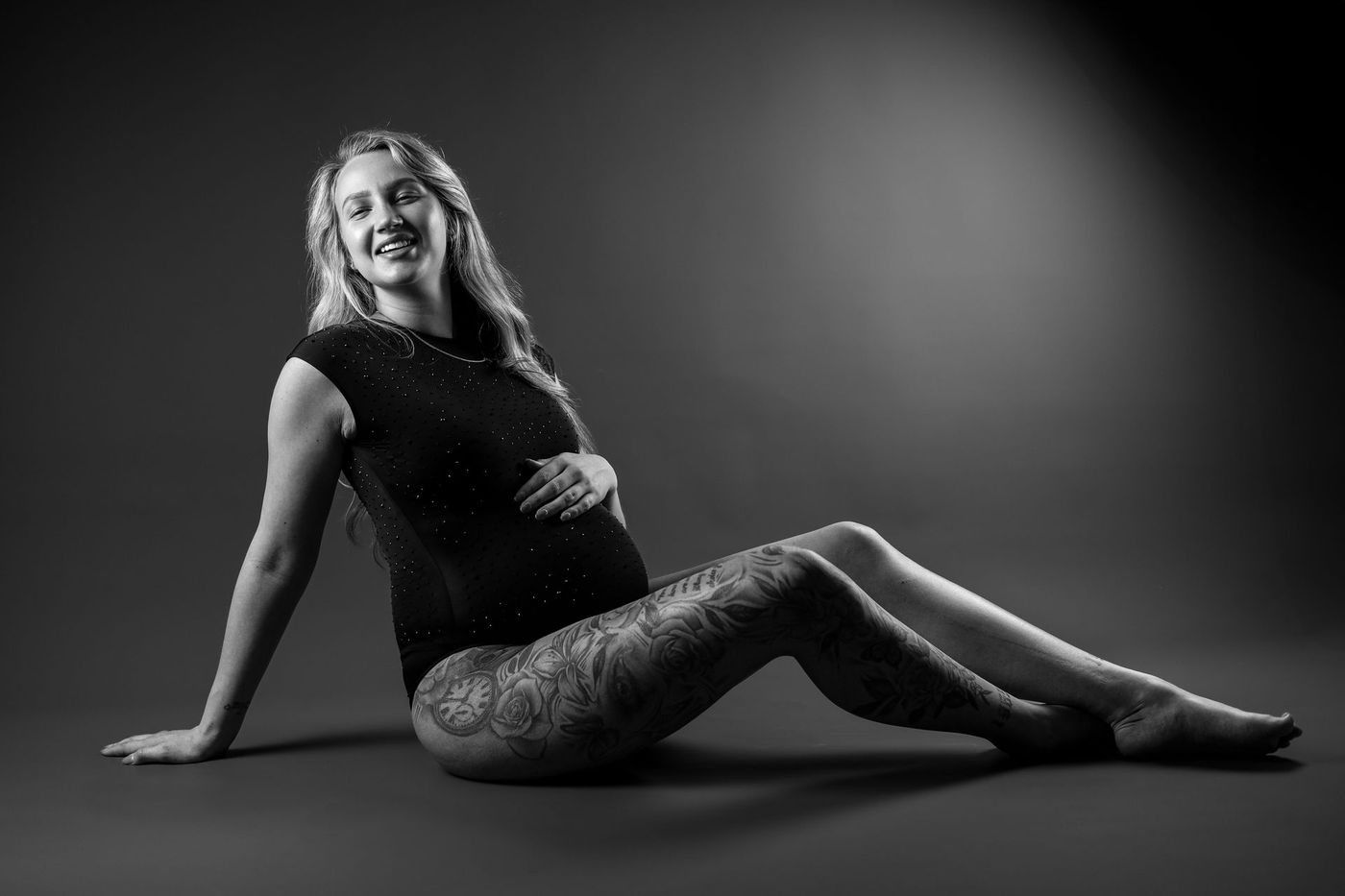 Vrouw ligt in een zwarte jurk op de vloer van een studio en lacht tegen een donkere, verlopende achtergrond.