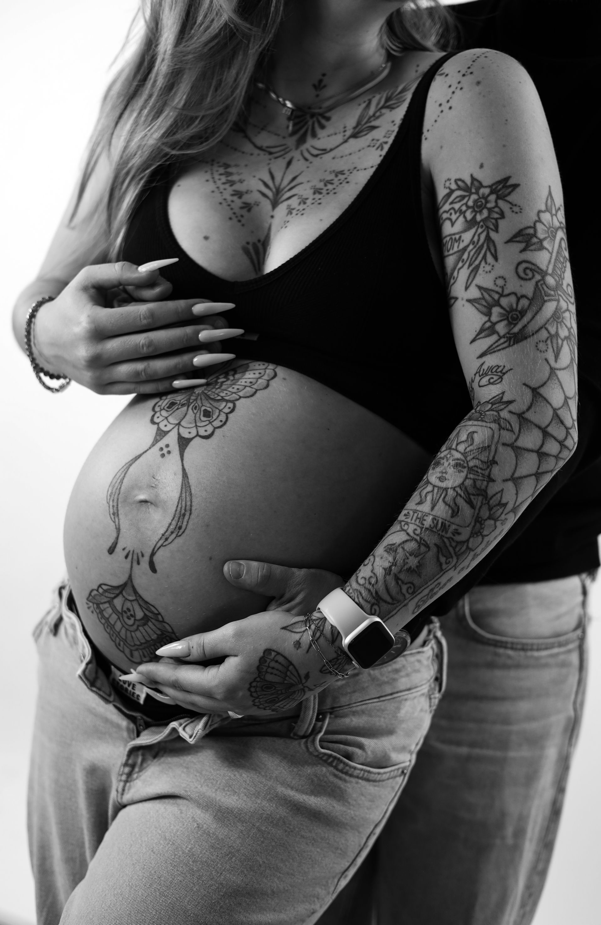 Zwart-witfoto van een zwangere vrouw met getatoeëerde armen die haar buik vasthoudt. Zwangerschap fotograaf.
