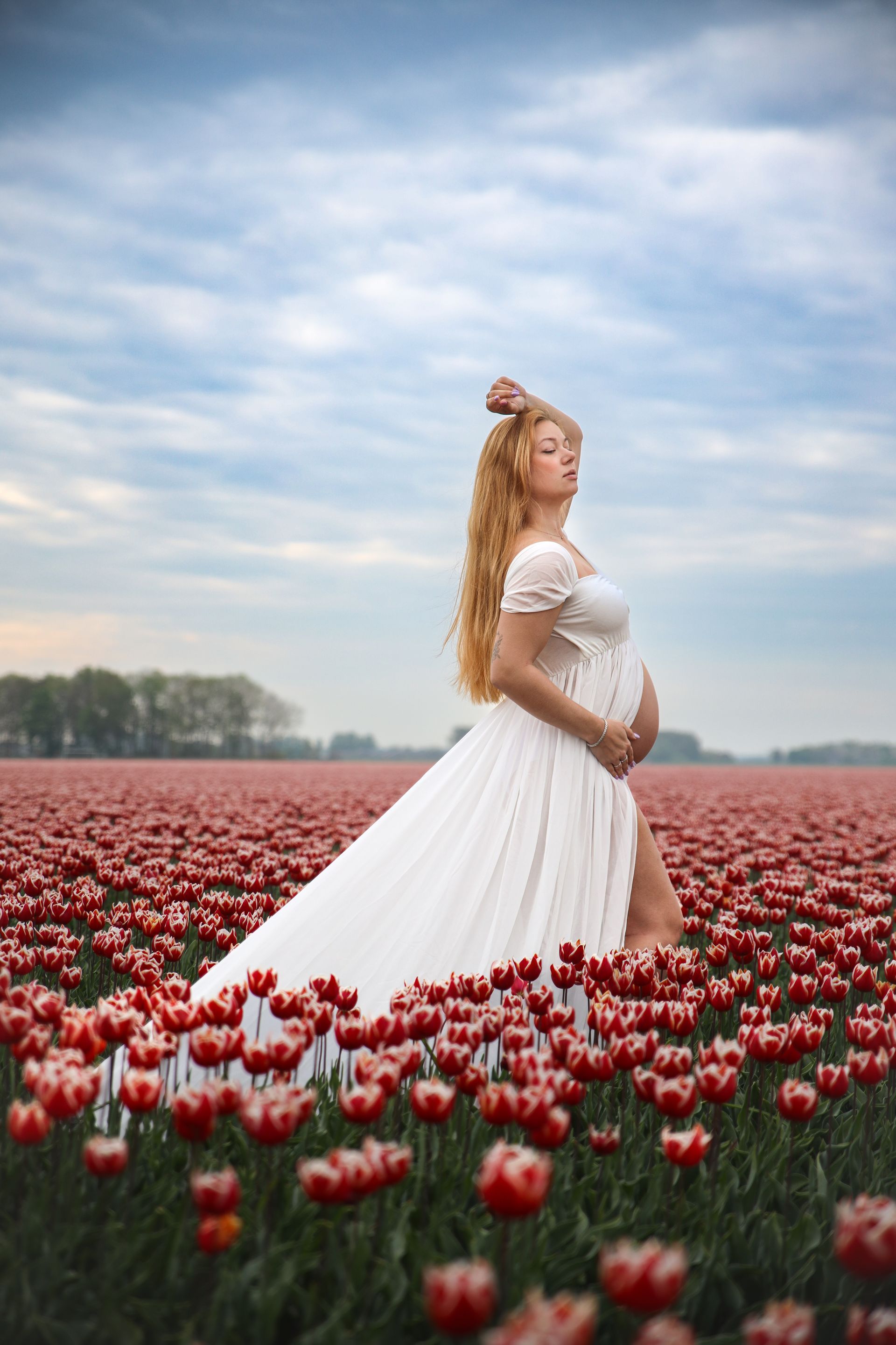 Een zwangere vrouw in een witte jurk staat in een veld met rode tulpen onder een bewolkte hemel. Goedkope zwangerschapsshoot.