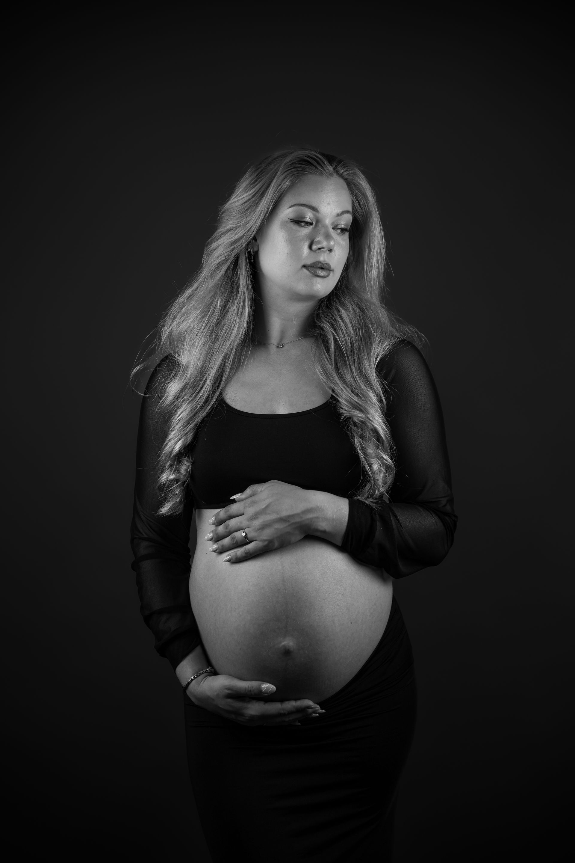 Een zwangere vrouw in een zwarte jurk houdt zachtjes haar buik vast tegen een donkere achtergrond. Zwangerschapsshoot.