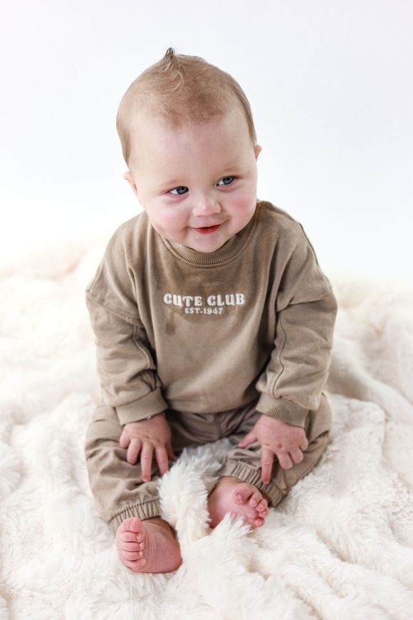 Een lachende baby in een beige shirtje zit op een zachte witte deken. Kinderfotografie. Kindershoot. Fotoshoot met kinderen.