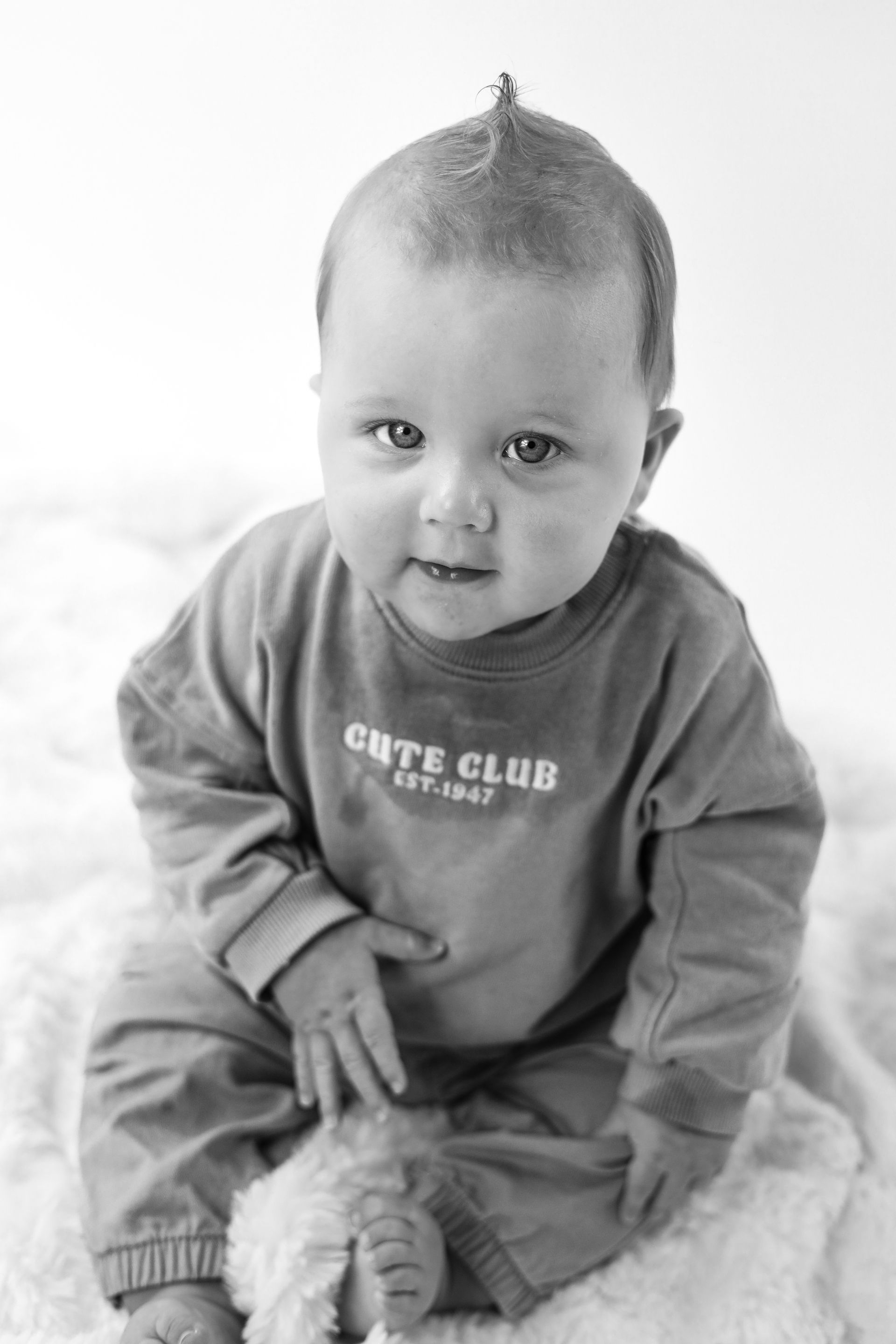De baby zit op een dekentje, draagt ​​een trui en een broek, en kijkt met een lichte glimlach naar de camera. Kinderfotoshoot