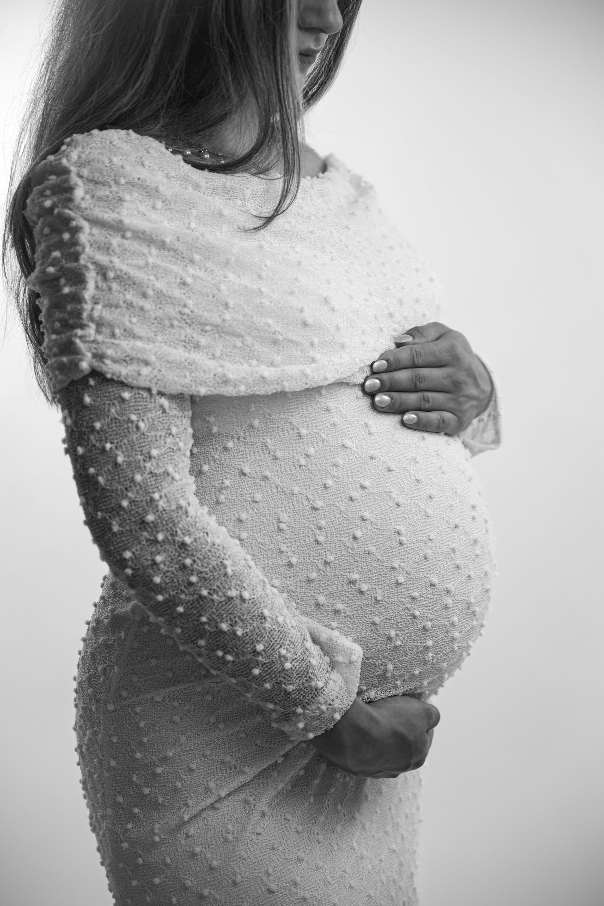 Een zwangere vrouw in een witte jurk houdt haar buik vast tegen een effen achtergrond. Zwangerschap fotograaf Voorne Putten.