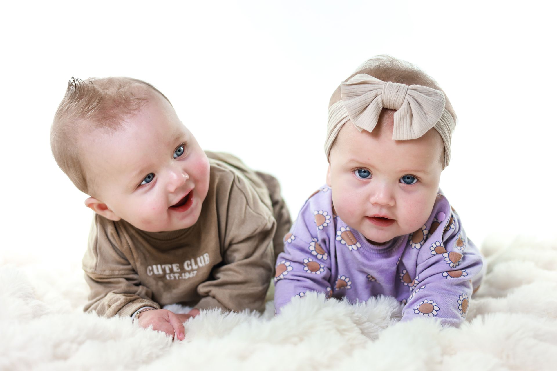 Twee baby's liggen op een zachte witte deken, gekleed in knusse outfits, en kijken naar de camera. Kinderfotograaf Zuidland.