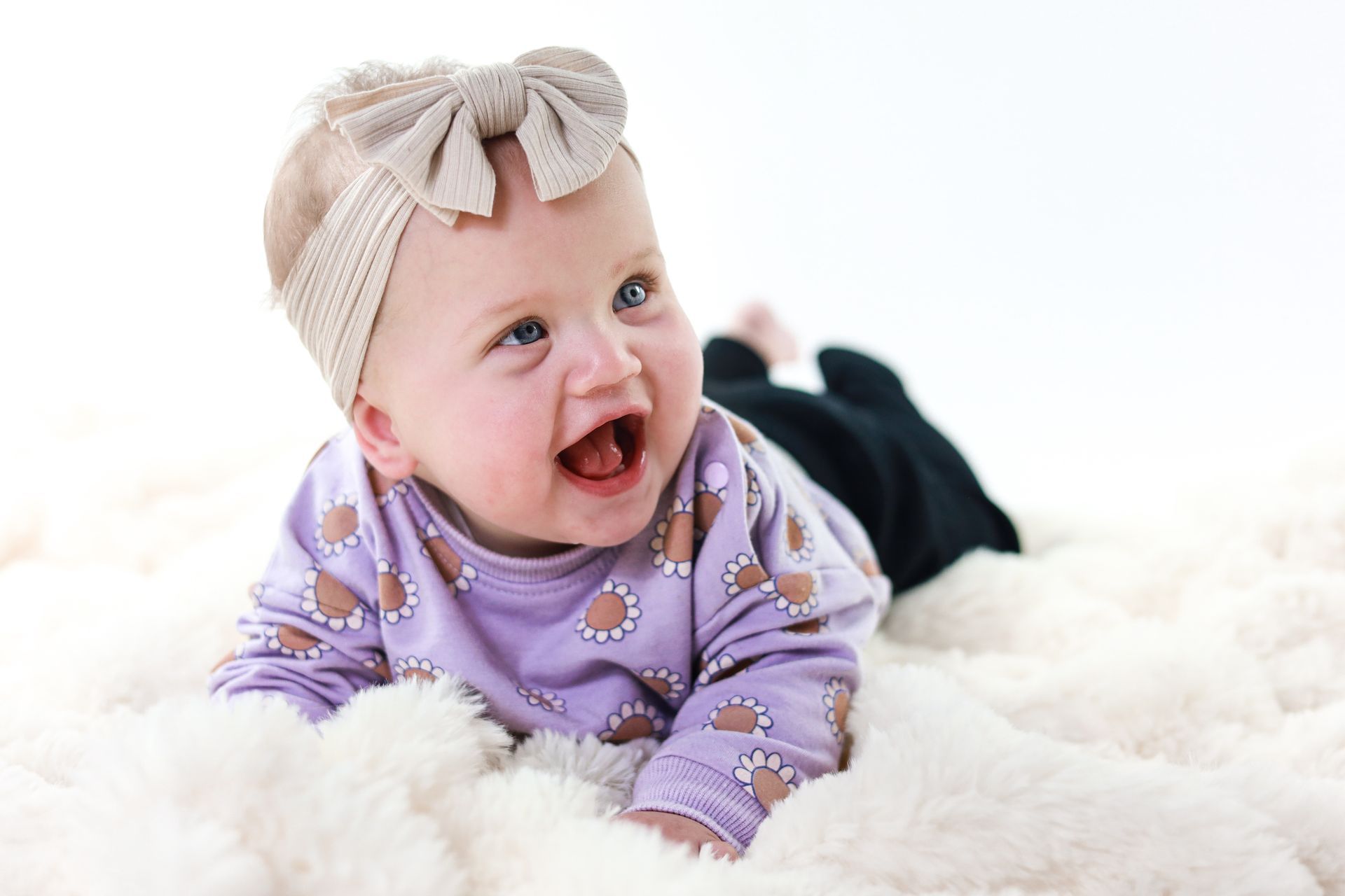 Een lachende baby in een paarse outfit en beige haarband ligt op een witte, pluizige deken. Kinderfotograaf. Babyfotoshoot.