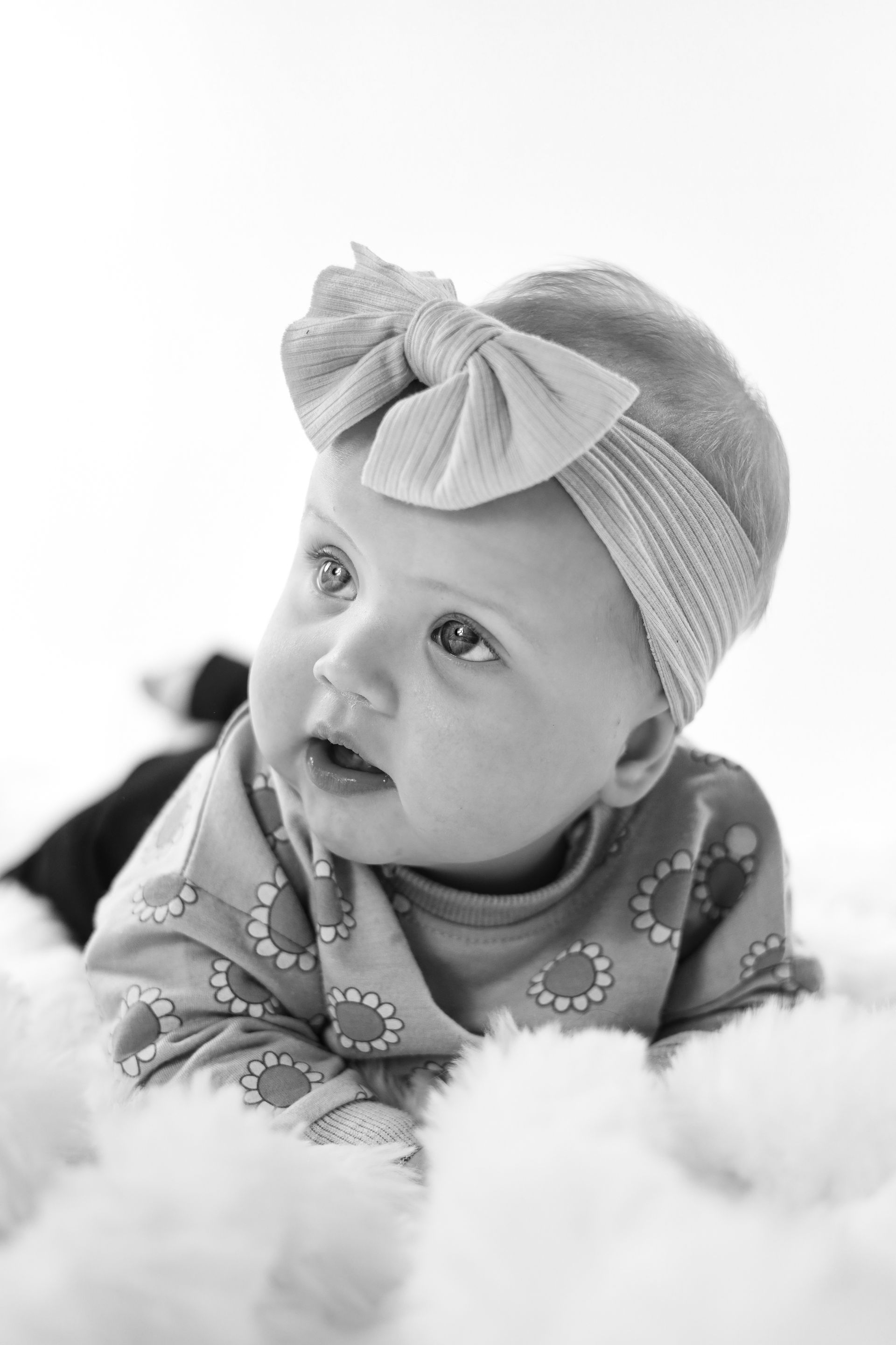 Een baby met een strik in haar haarband ligt op een zachte deken en kijkt opzij. Peuter fotoshoot. Kinderfoto.