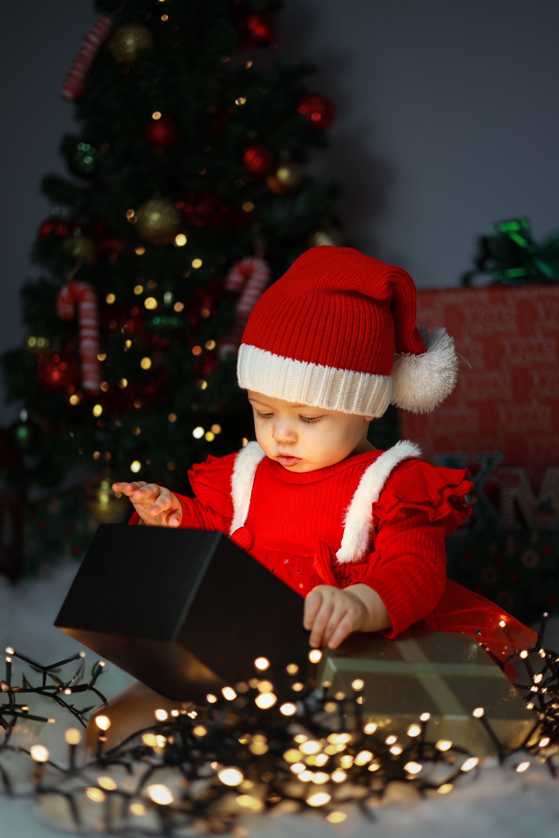 Een baby in een rood kerstmanpakje opent een cadeautje bij kerstverlichting en een versierde kerstboom. Kerst fotoshoot.
