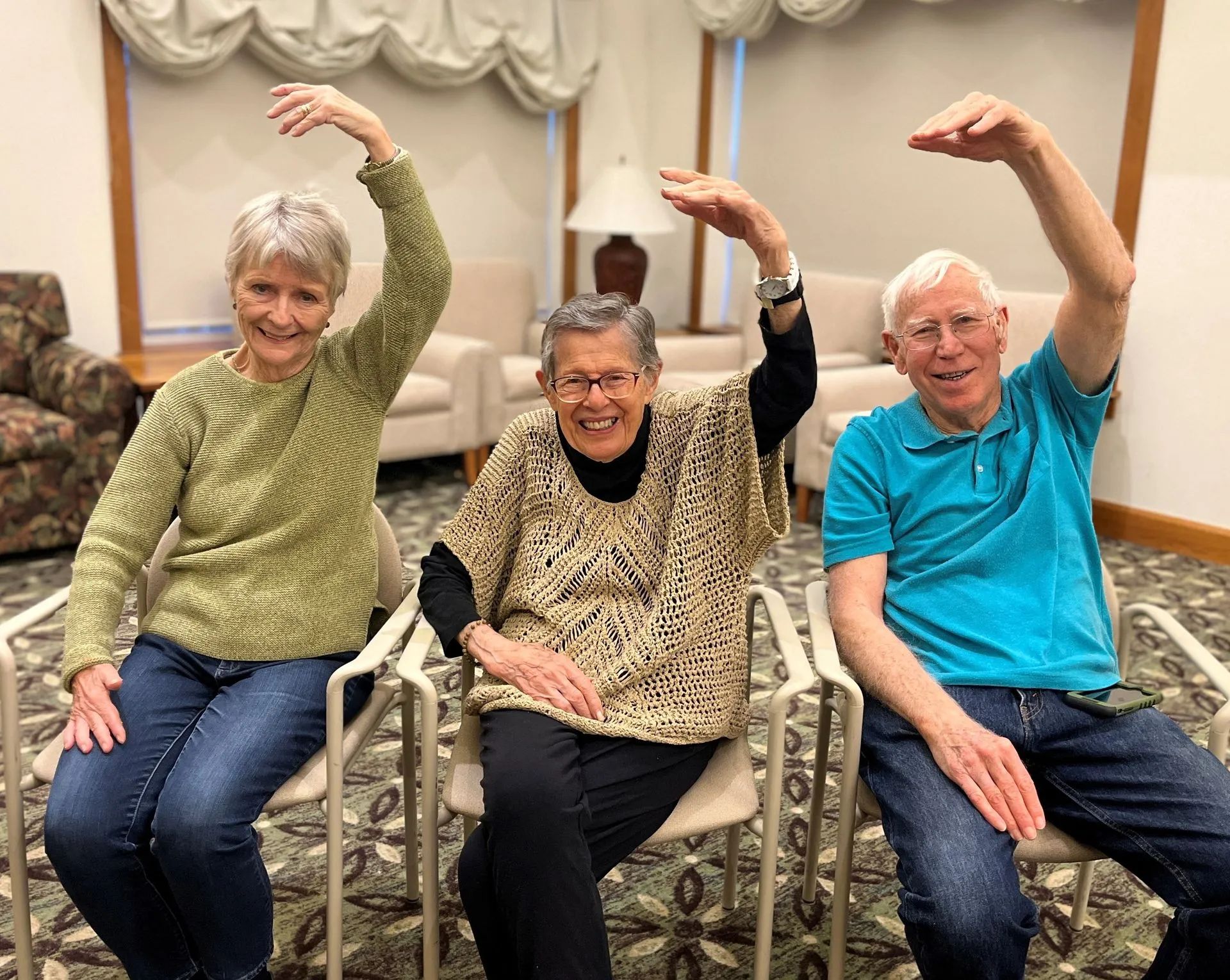 Three people seated, arms arched overhead, smiling. Indoors, chairs, beige and green tones.