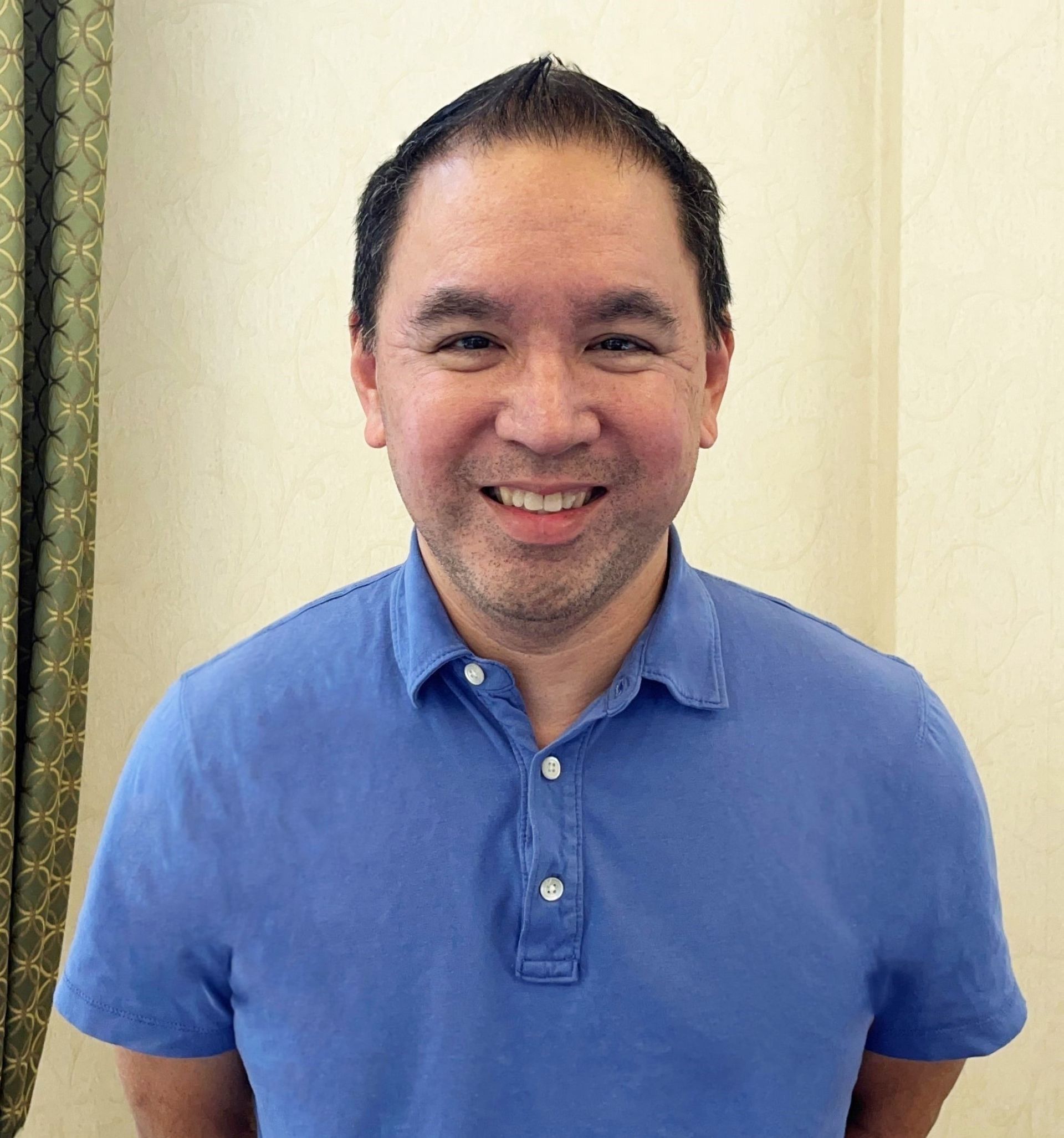 Man in blue polo shirt smiling, standing indoors against a cream wall.