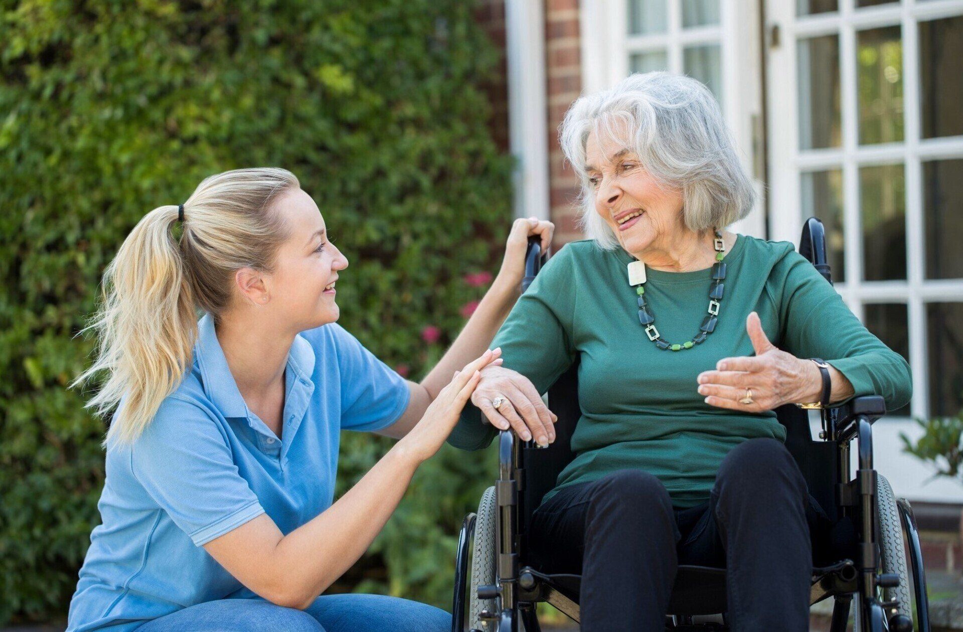 Woman in wheelchair talking to caregiver outside a house, both smiling.