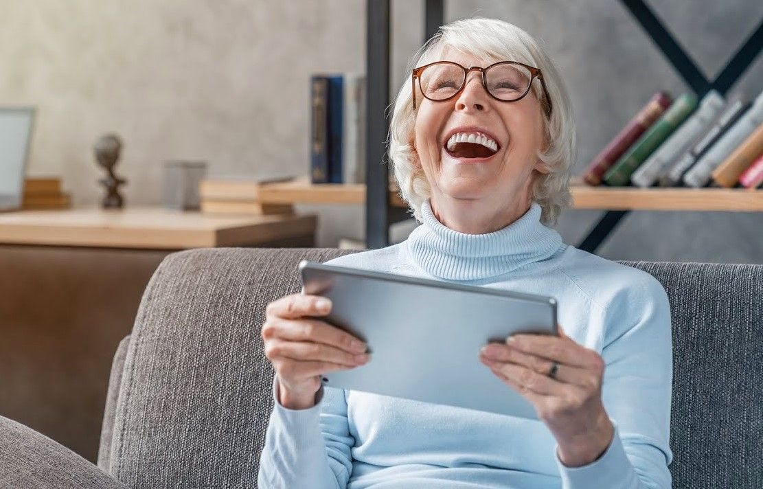 Woman laughing while holding tablet. Sitting on a sofa, wearing glasses and a blue sweater.