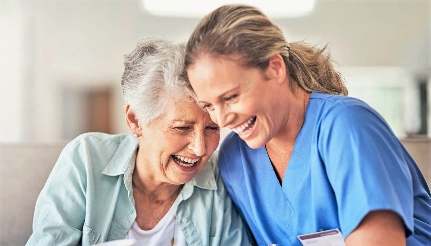 A senior woman and a care worker laughing, close together, in a brightly lit setting.