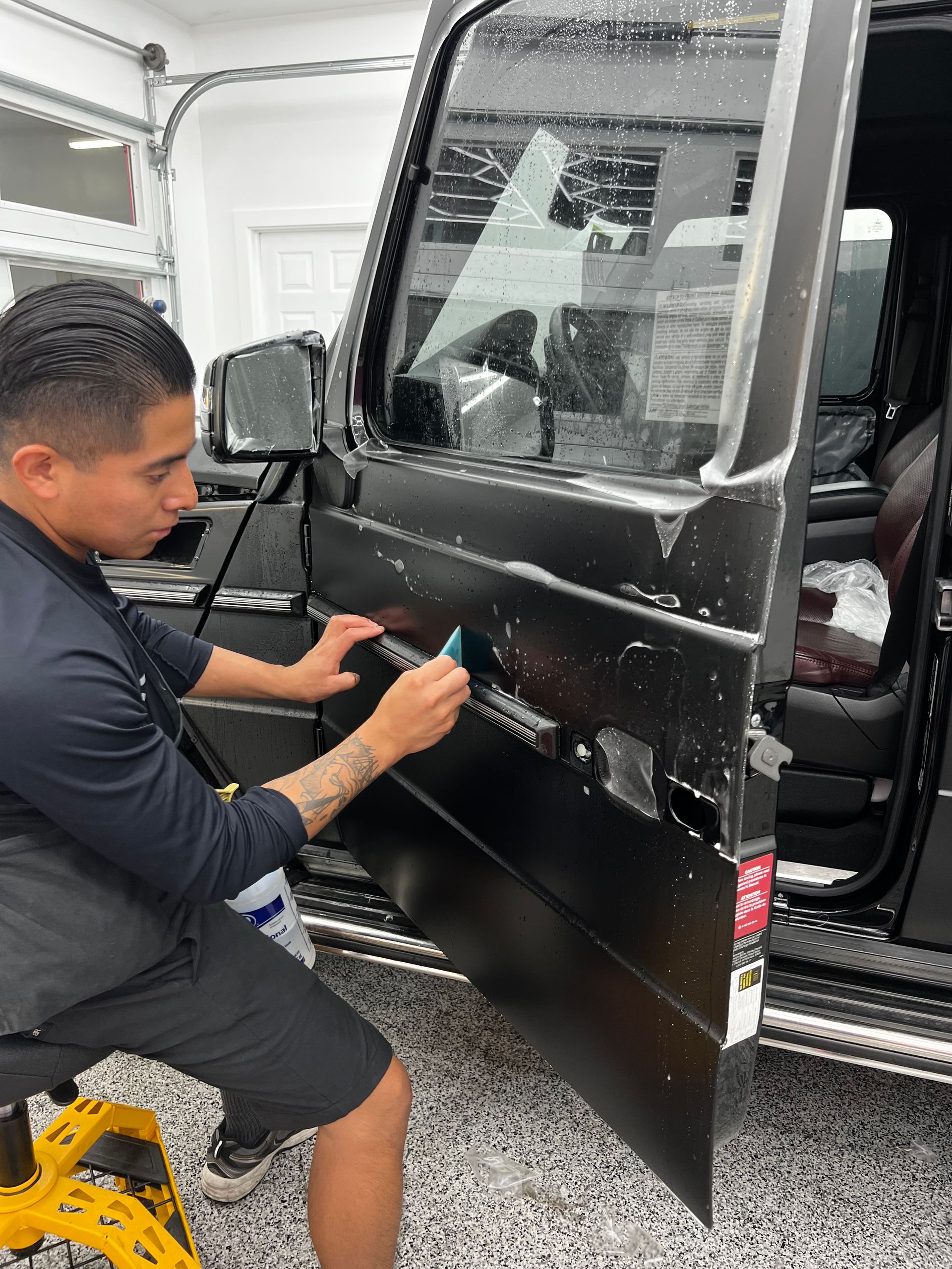 A technician applies a protective film to the black door of a vehicle in a bright garage setting.