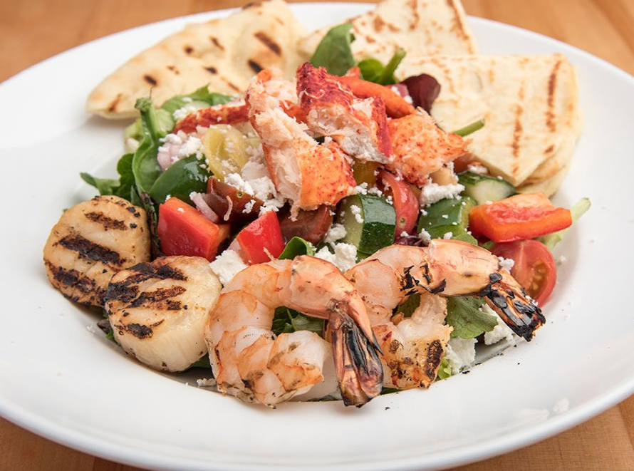 A white plate topped with a salad with shrimp , scallops , tomatoes and pita bread.