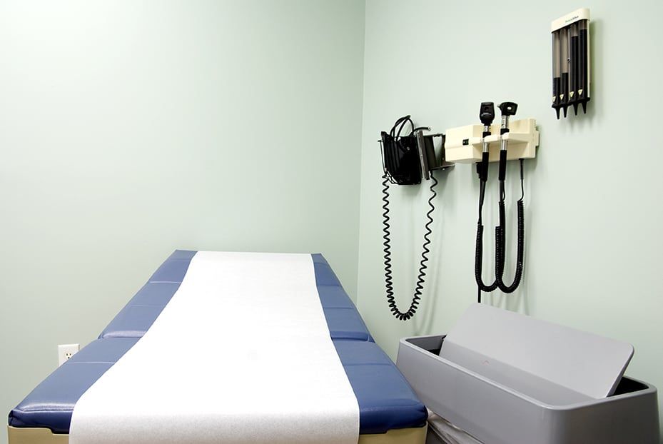 An examination room with a blue exam table, medical diagnostic equipment wall-mounted above, and a gray waste bin nearby.