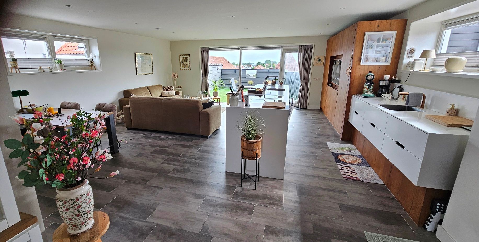 Lichte woonkamer met moderne keuken, tegelvloer, banken en eethoek en toegang tot het dakterras. 