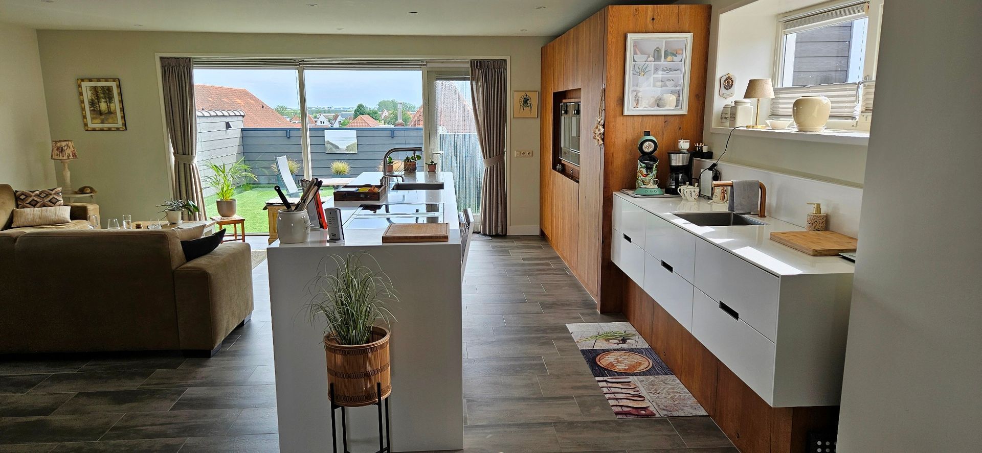 Lichte woonkamer met moderne keuken, tegelvloer, banken en eethoek en toegang tot het dakterras. 