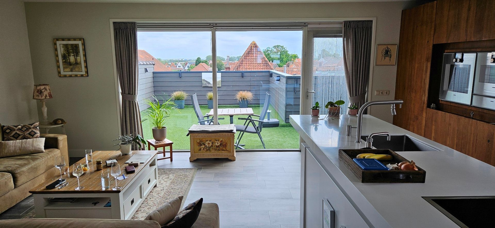 Lichte woonkamer met moderne keuken, tegelvloer, banken en eethoek en toegang tot het dakterras. 