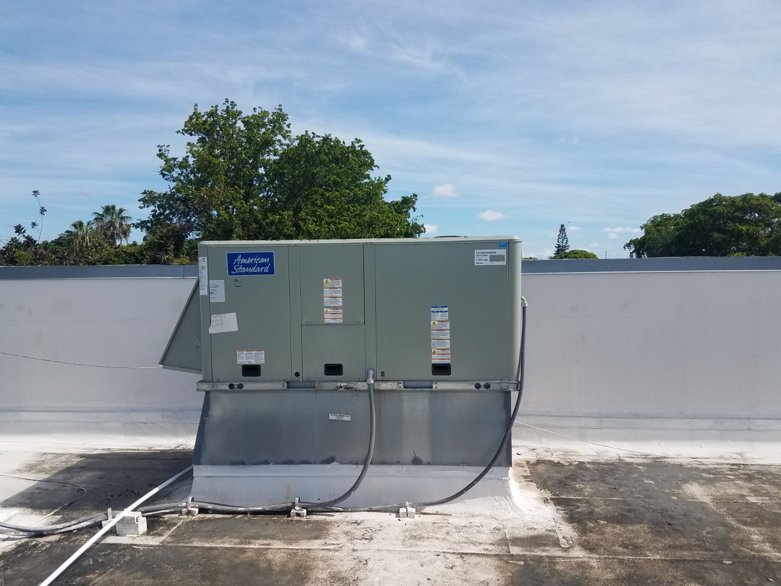A rooftop air conditioning unit on a flat white roof, with a tree and blue sky in the background.