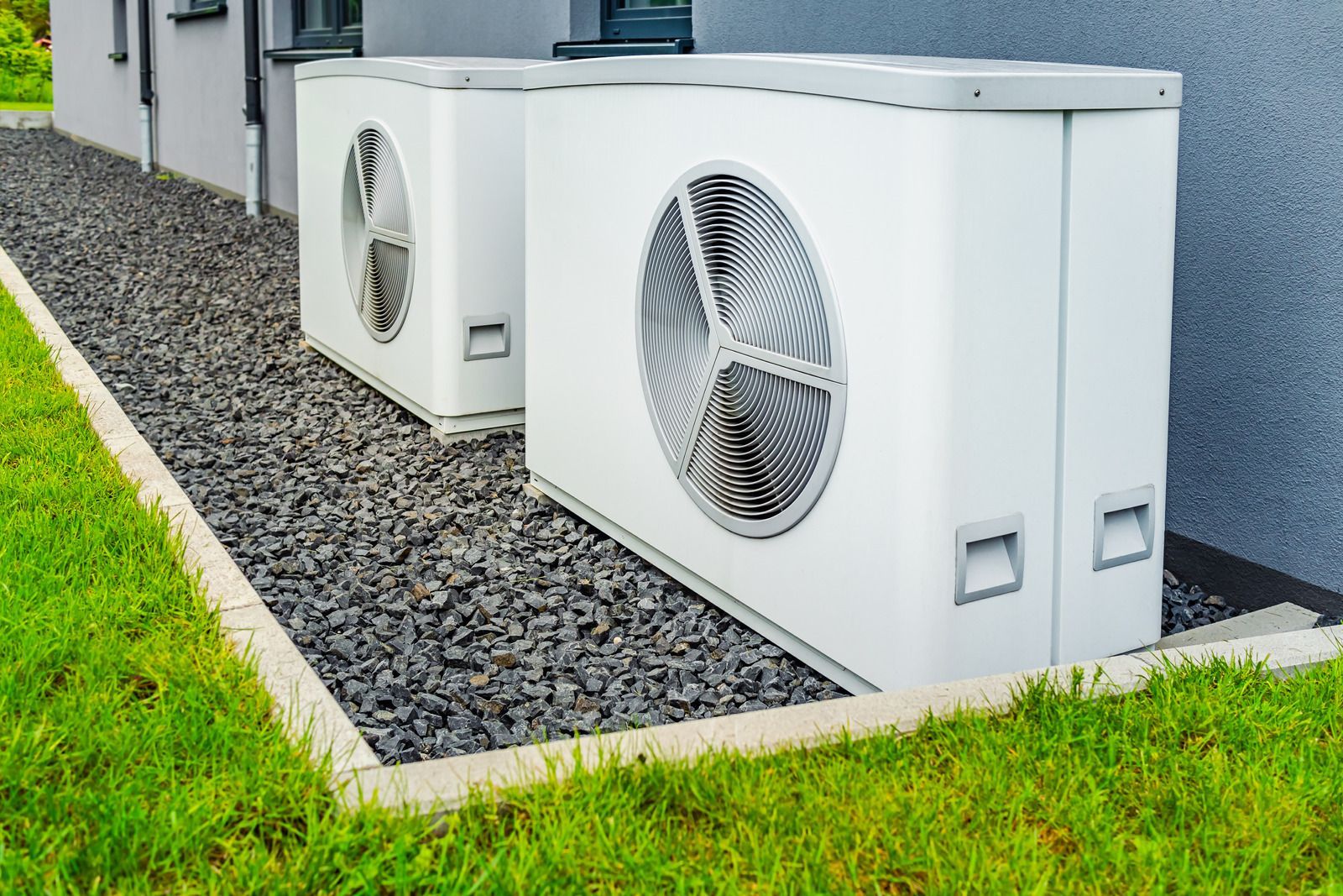 Two white heat pump units on a bed of rocks next to a building and green grass.