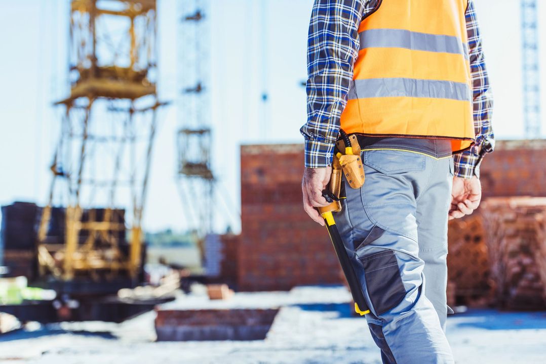 Un operaio edile con un giubbotto di sicurezza arancione cammina in un cantiere con delle gru sullo sfondo.
