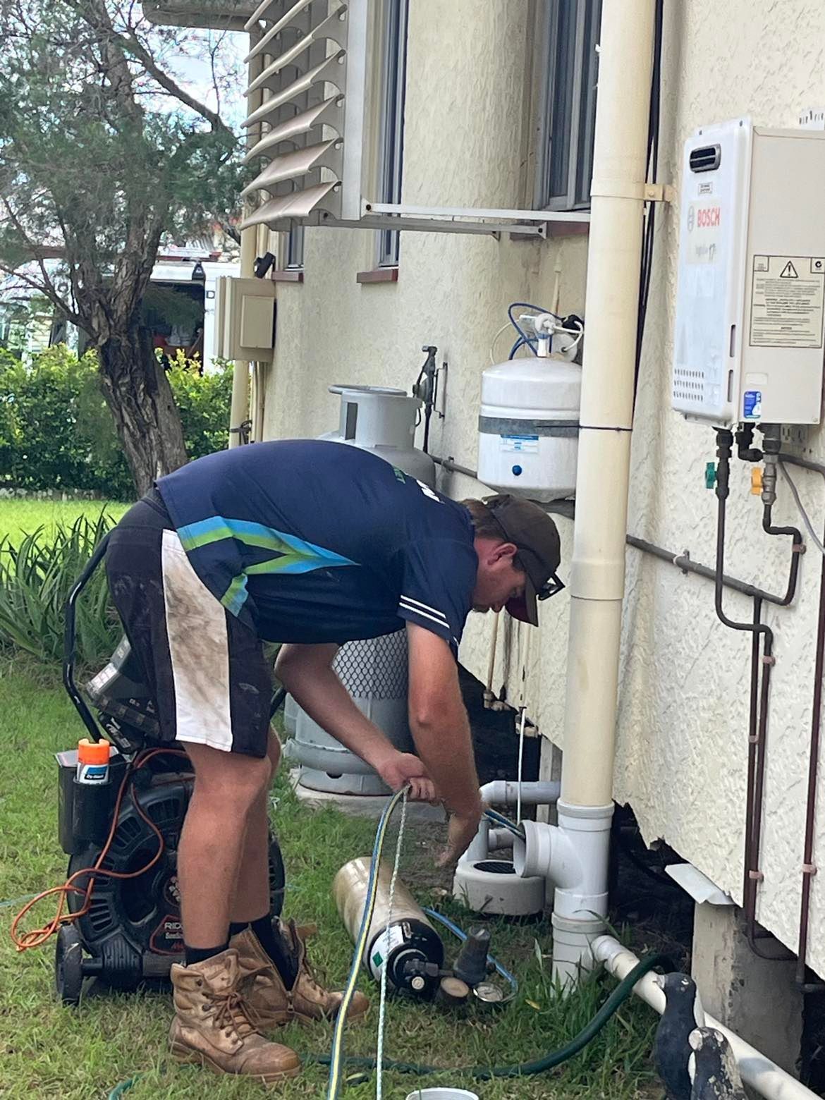 Plumber working on pipes outside a house. He is wearing a blue shirt and shorts, in a yard. — Nuflow Wide Bay in South Kolan, QLD