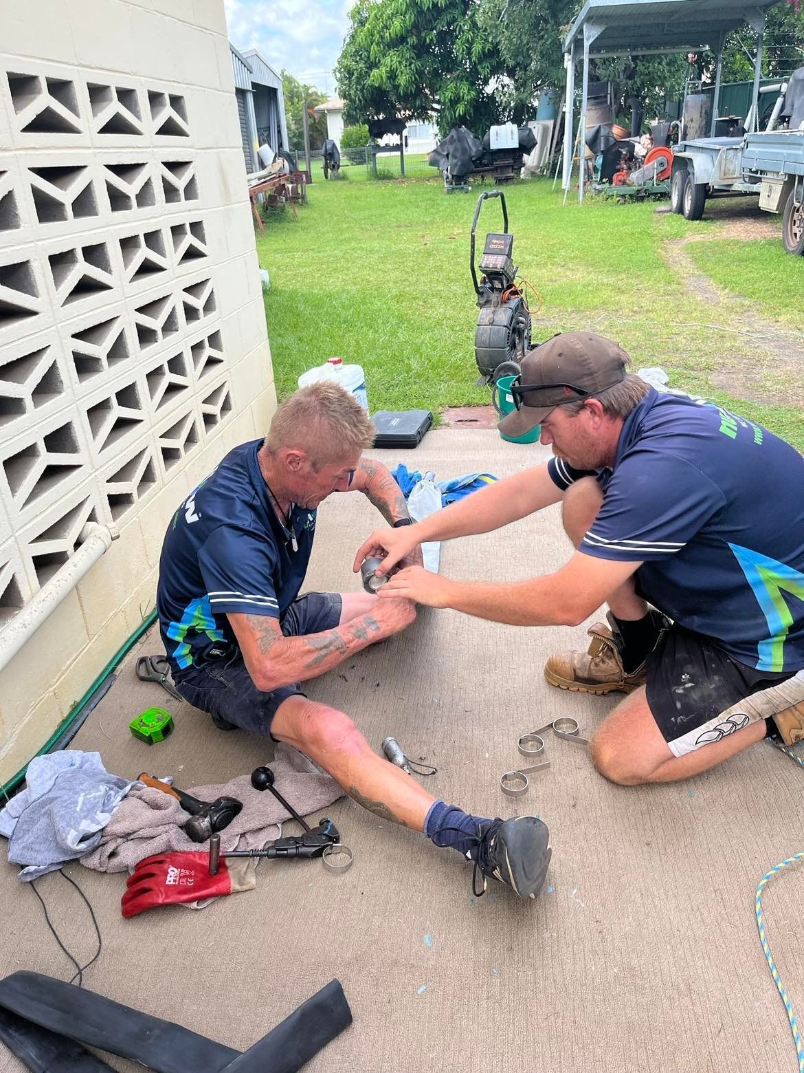 Two men working on equipment outdoors; one examines the other's arm, possibly a wound, near a building with a patterned wall. — Nuflow Wide Bay in Maryborough, QLD