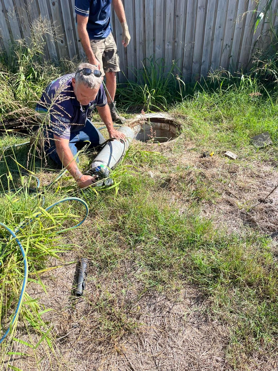 Two men near a manhole in a grassy area; one kneels, holding a pipe, next to the open manhole. — Nuflow Wide Bay in Childers, QLD