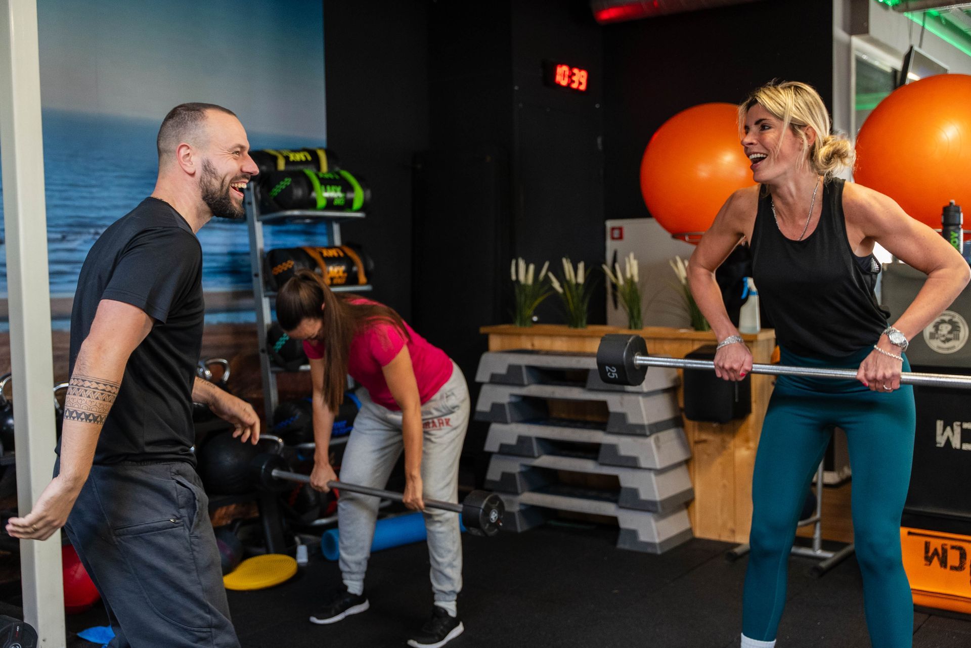 Mensen lachen in een sportschool: twee vrouwen en een man tillen gewichten. Oranje fitnessballen zijn zichtbaar.