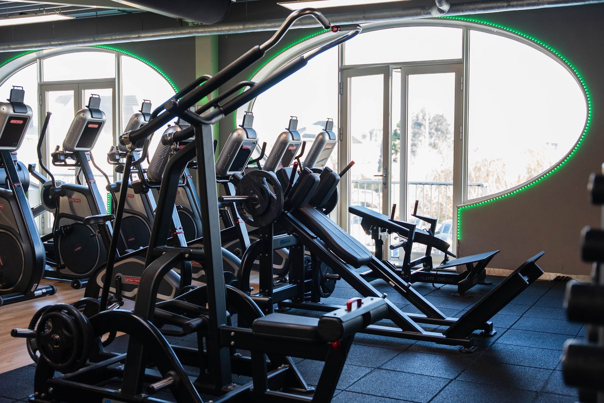 Interieur van een sportschool met fitnessapparaten, waaronder een leg press, vlakbij een groot raam.
