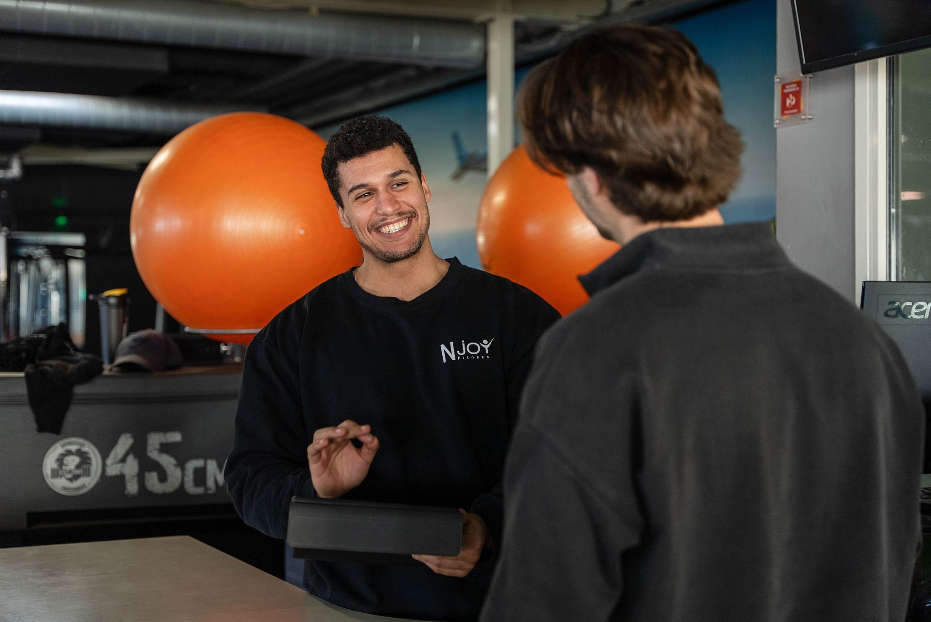 Een medewerker van de sportschool glimlacht, gebaart met een tablet en praat met een klant aan de balie, met oranje fitnessballen op de achtergrond.