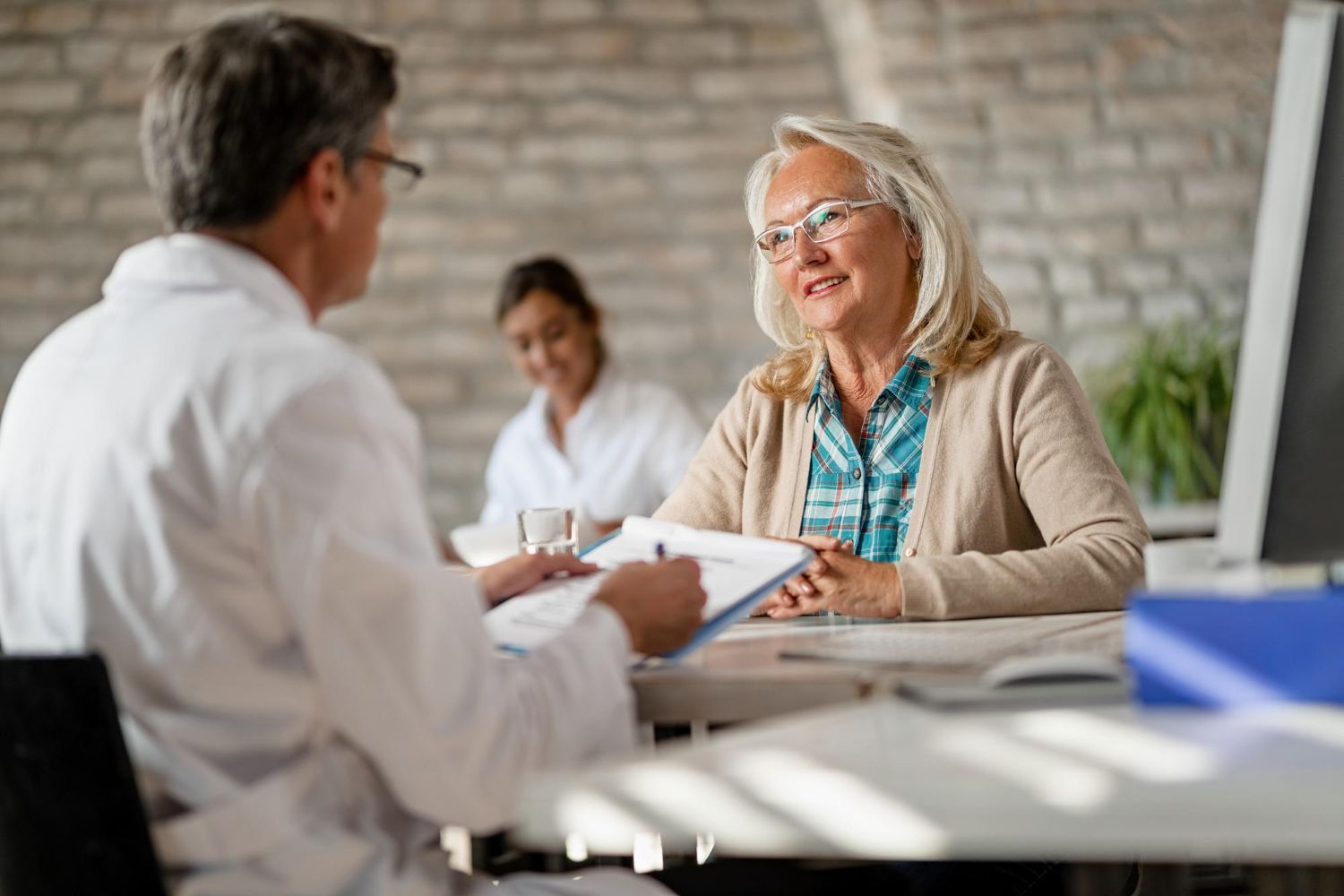 Two people in a bright office discussing documents across a table, with another person blurred in the background.