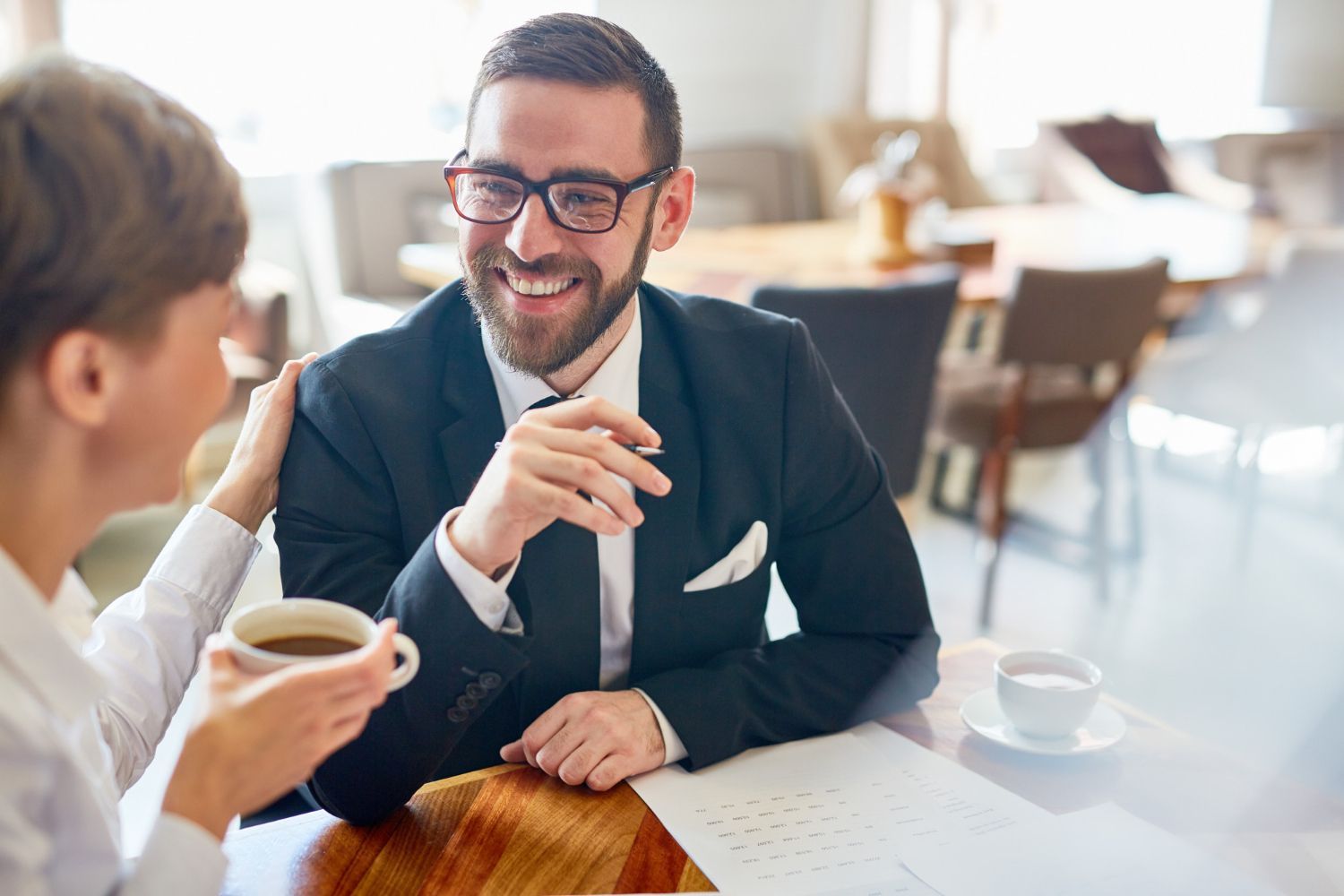 Two people talking over coffee in a bright café, one smiling in a dark suit