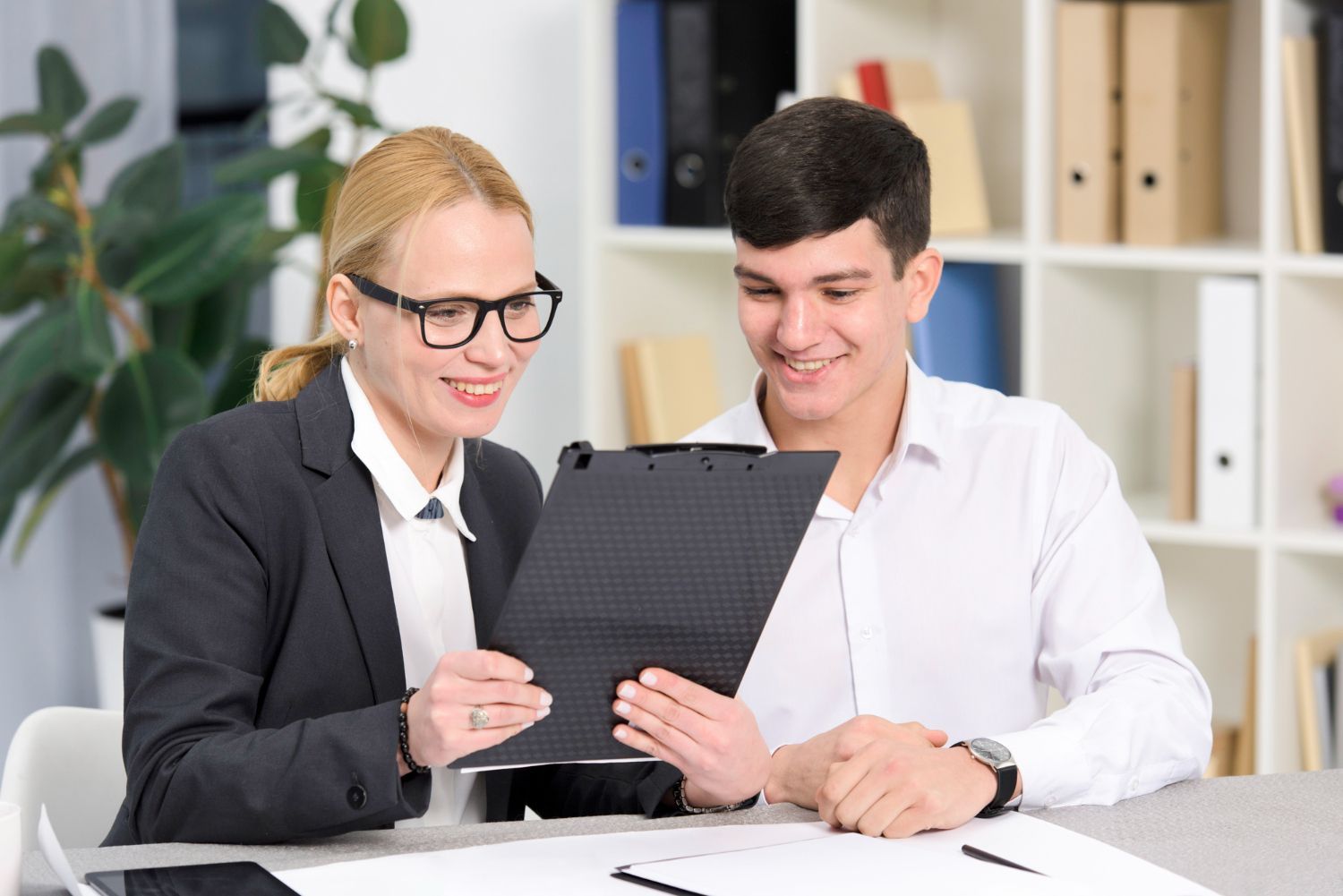 Two coworkers reviewing a clipboard at a desk in an office, smiling and discussing notes