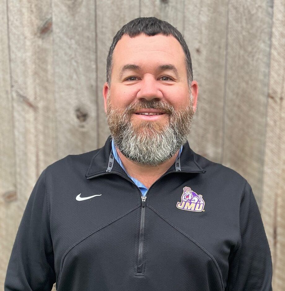 Smiling man in a black pullover with a logo, standing in front of a wooden wall.