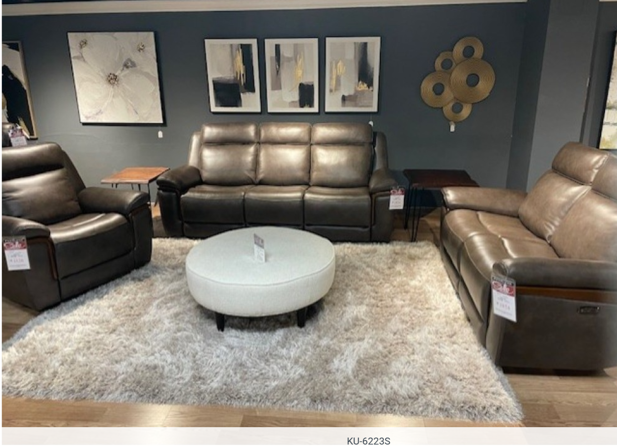 Living room furniture set: brown leather sofas and recliner, round white ottoman, beige shaggy rug, artwork.