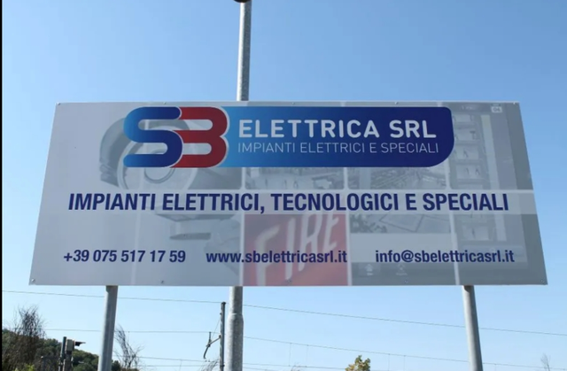 Cartellone pubblicitario per SB Elettrica SRL, azienda di impianti elettrici, con i recapiti su un'insegna bianca su sfondo blu.