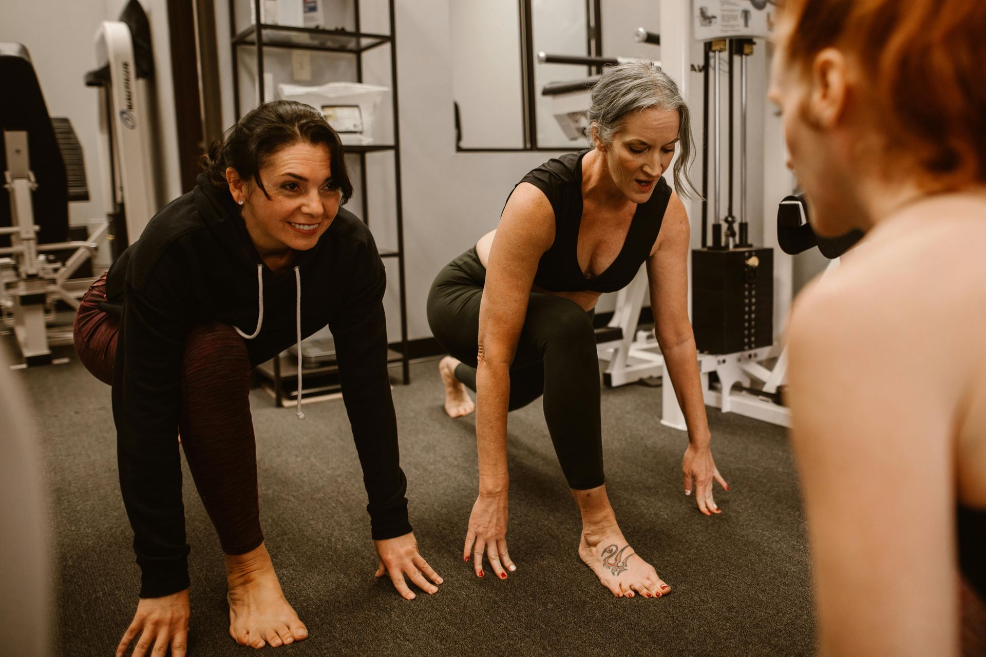Two women squatting in gym, hands on floor. One woman looking at another woman. Gym setting.