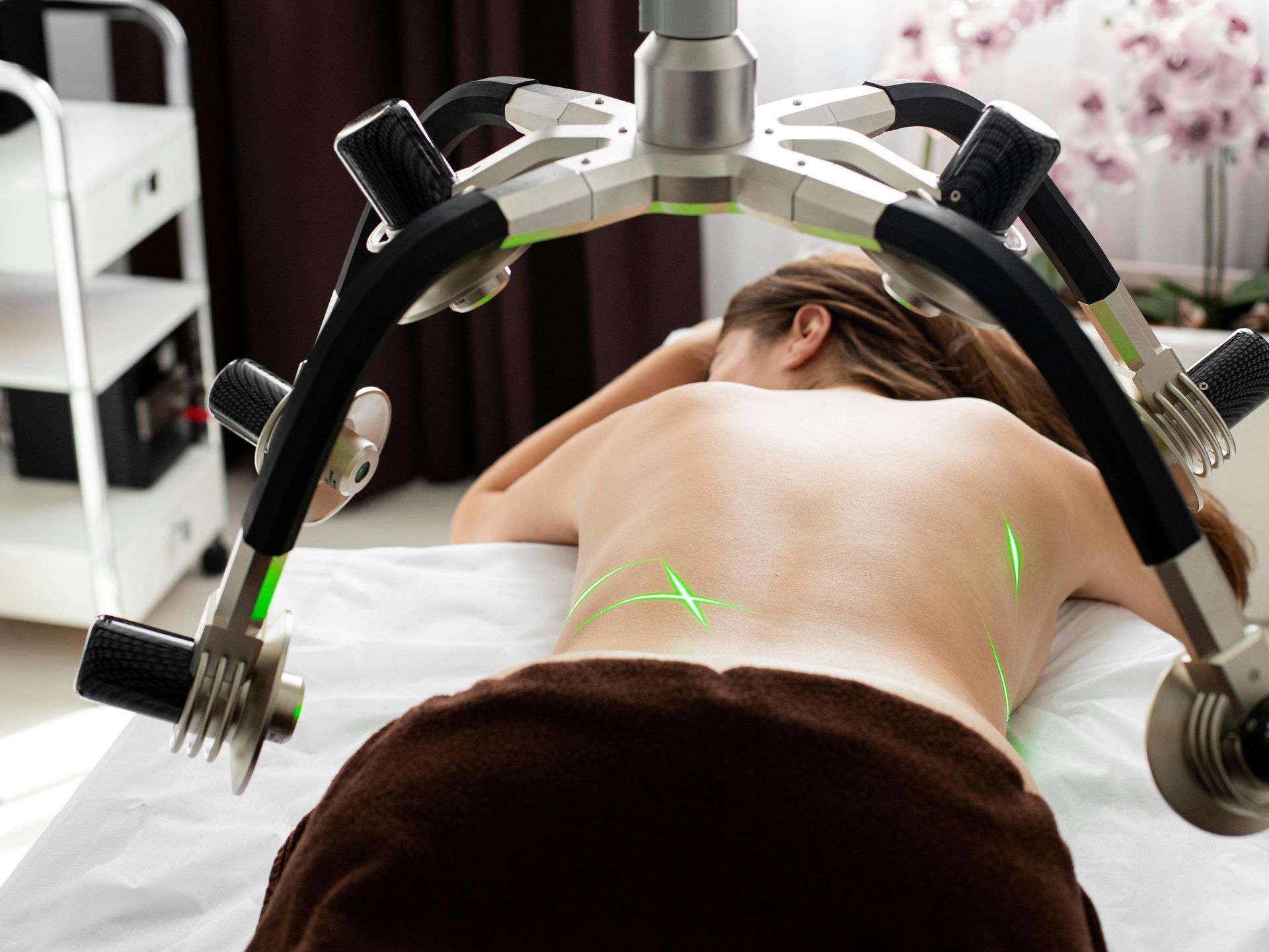 Woman receiving laser therapy on her back, lying face down on a massage table. Green laser beams are visible.