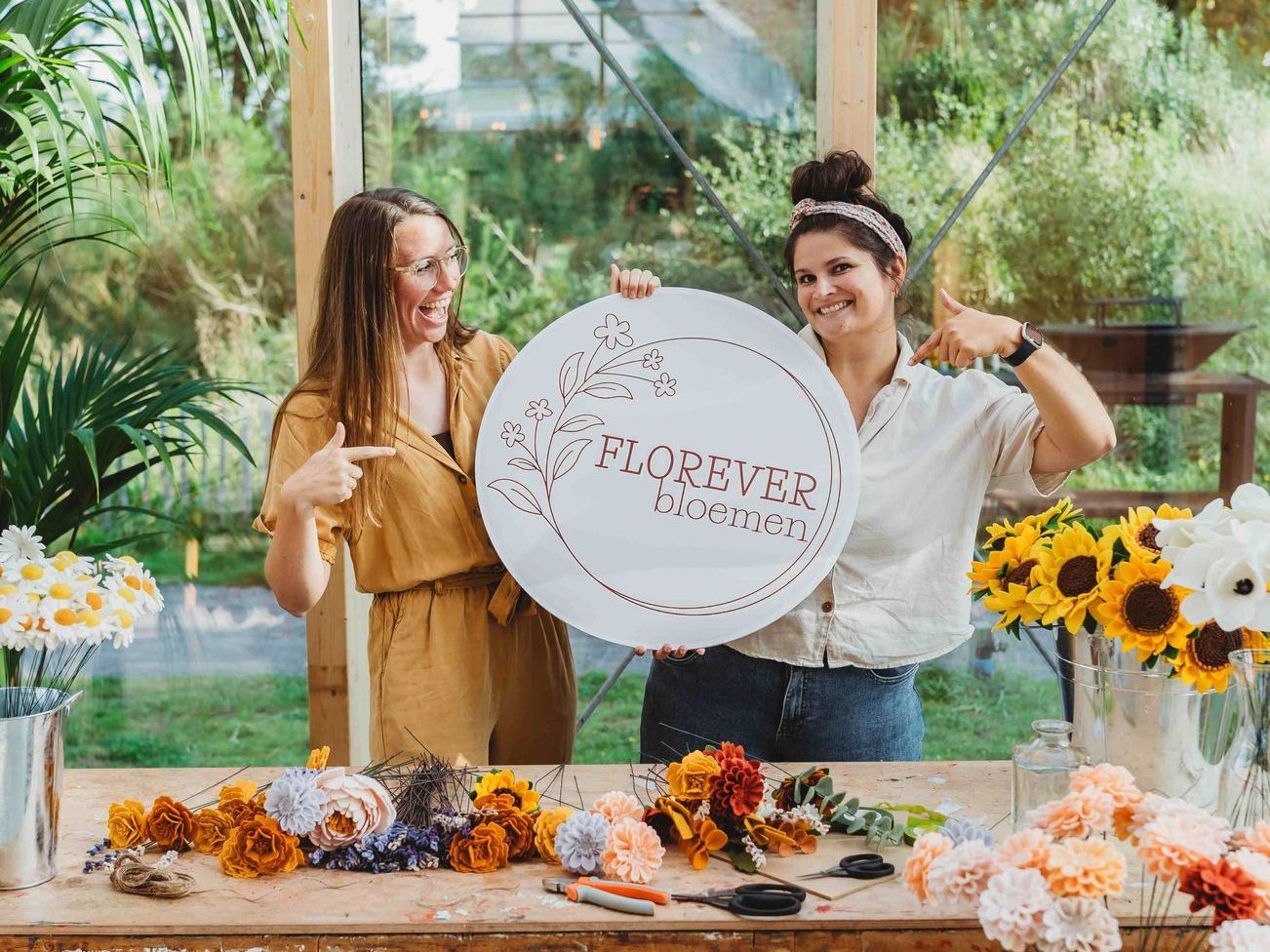 de ontwerpers staan ​​naast elkaar met een bord in de hand met daarop hun logo voor een tafel vol viltbloemen.