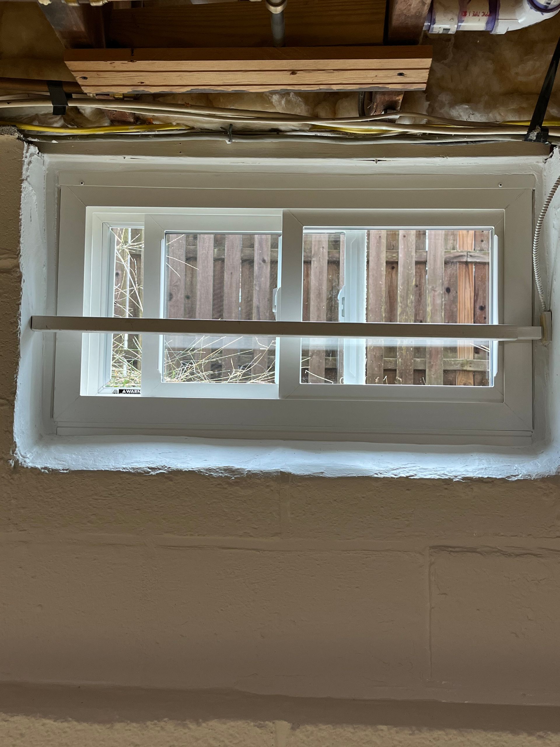 Small white basement window with horizontal bars set in a concrete wall, with exposed beams above.